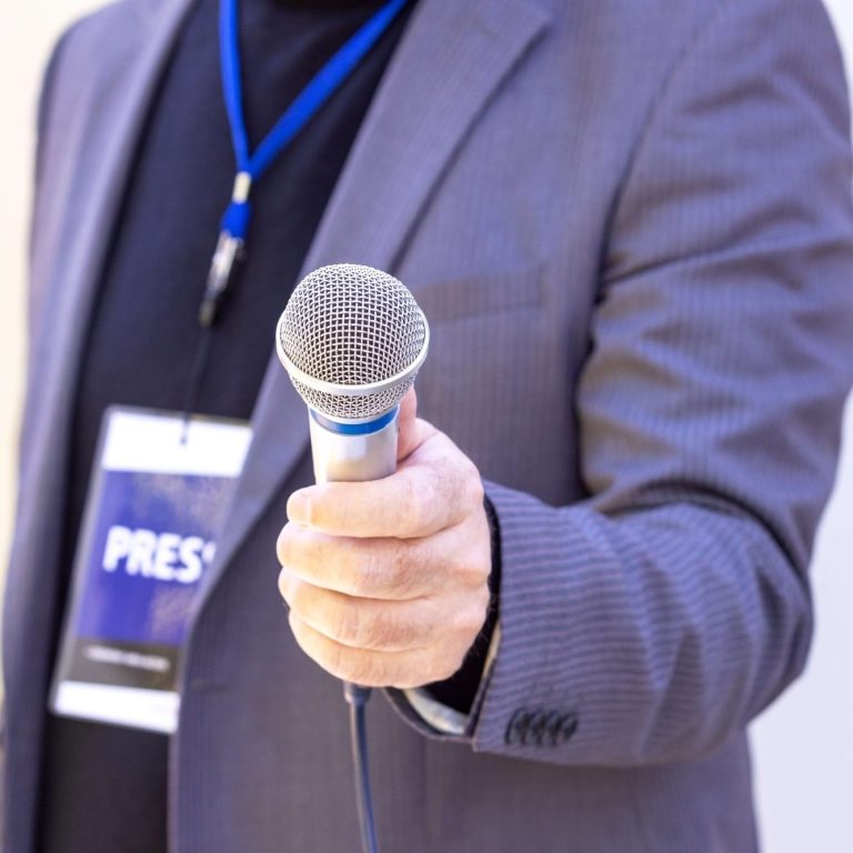 Journalist holding a microphone and a note pad A person holding a microphone, wearing a press badge and a blue lanyard.